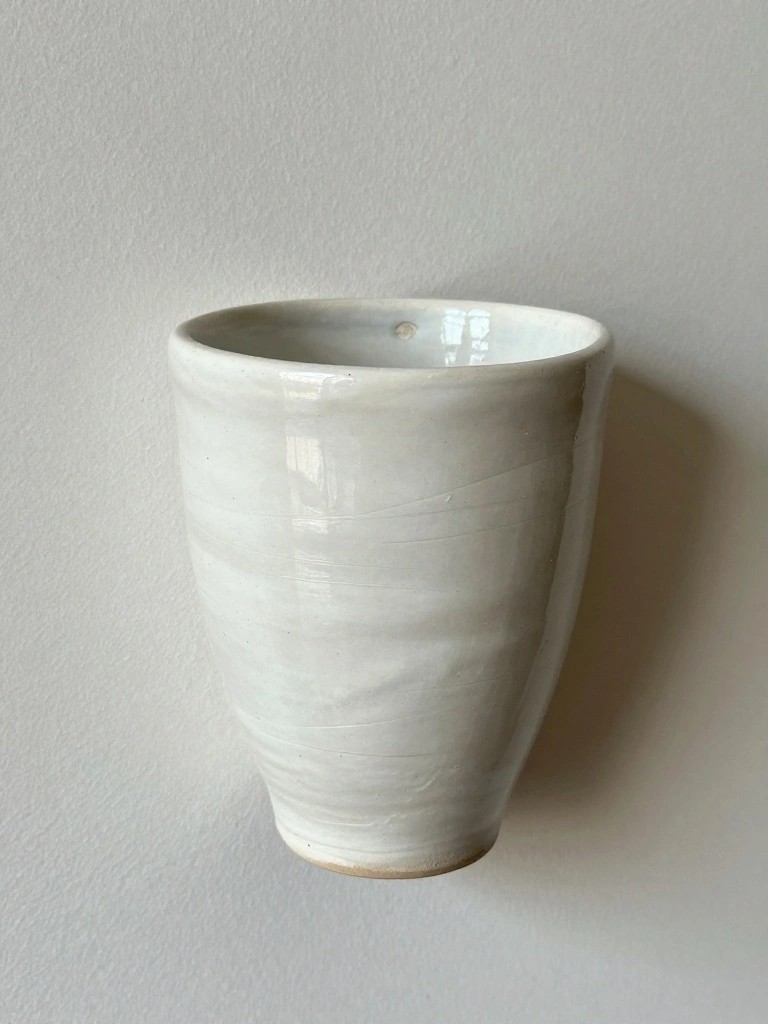 A white ceramic mug with glossy glaze highlights and a small rim loop hangs on a white wall.