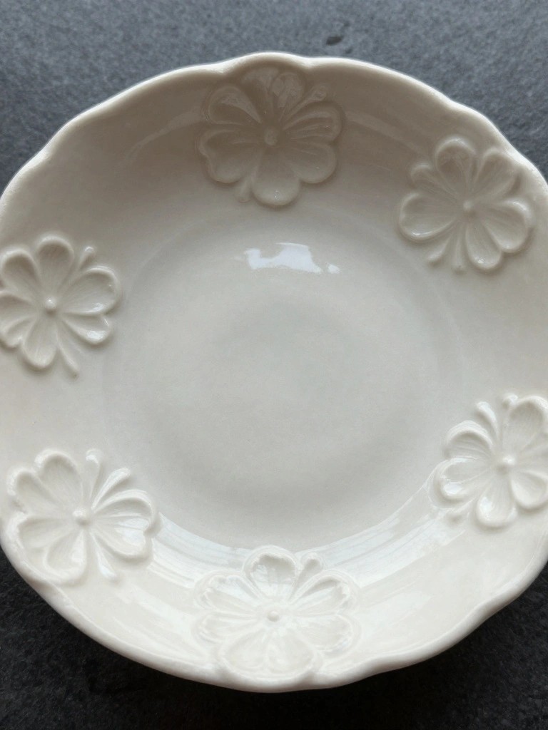 White ceramic shallow bowl with scalloped rim and five raised white flower motifs around the interior edge on a dark surface.
