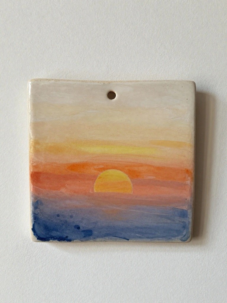 Square handmade ceramic tile with a painted gradient sunset over blue water and a hanging hole at the top.