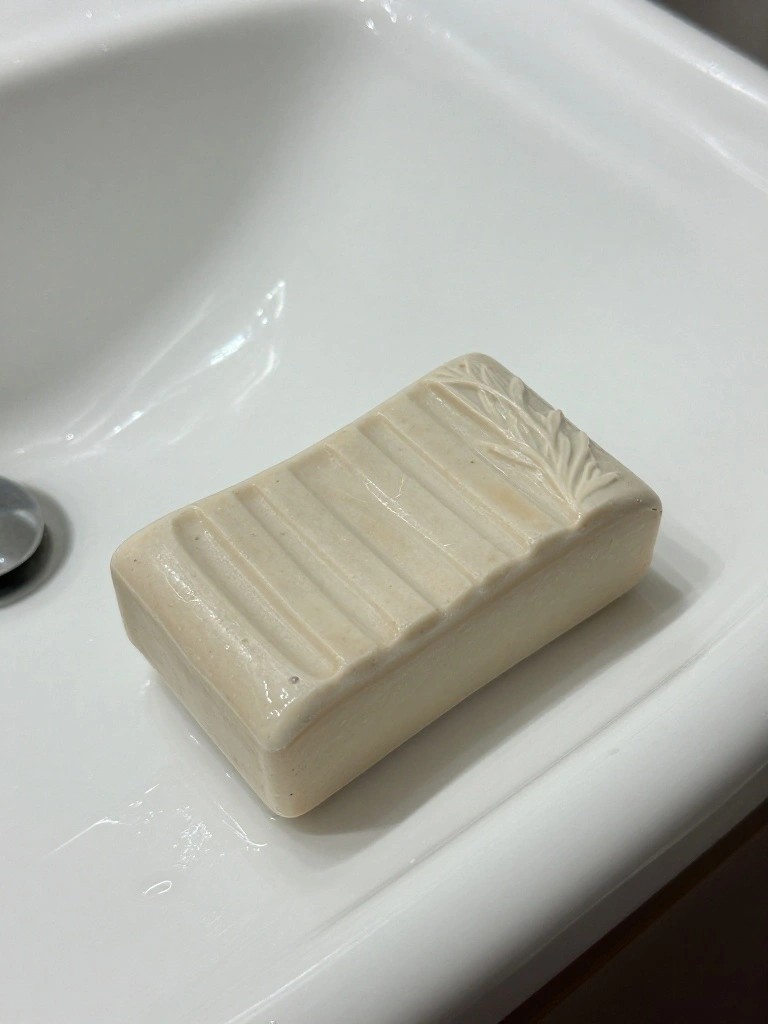 A handmade beige ceramic bar shaped like ridged soap with a leaf motif sits in a white porcelain sink next to the drain.