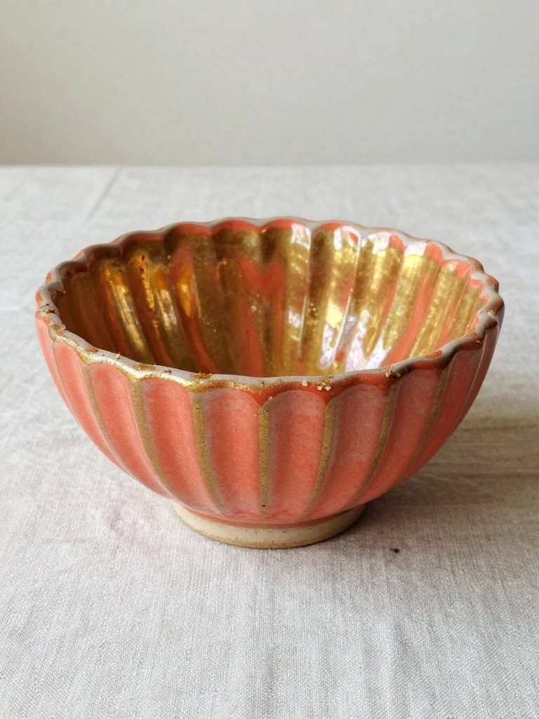 A small handmade ceramic bowl with fluted edges in coral glaze and gold ridges on a textured white surface.