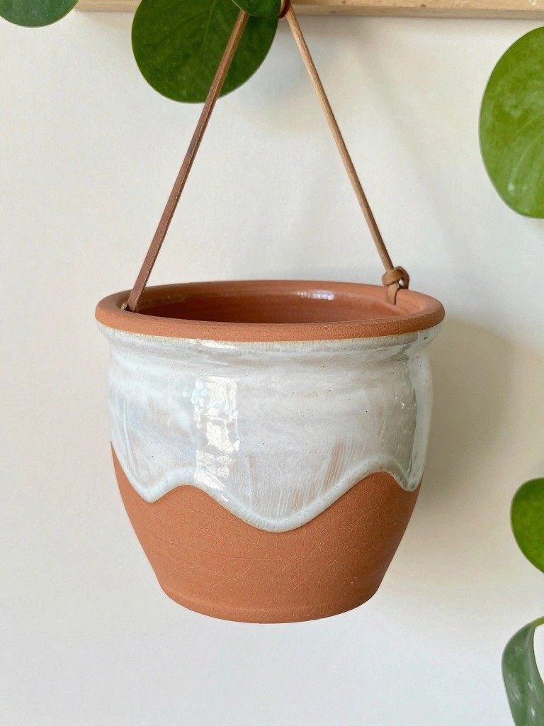 A handmade terracotta hanging planter with white wavy glaze on the upper half, suspended by brown ropes from a wooden beam with green plant leaves nearby.