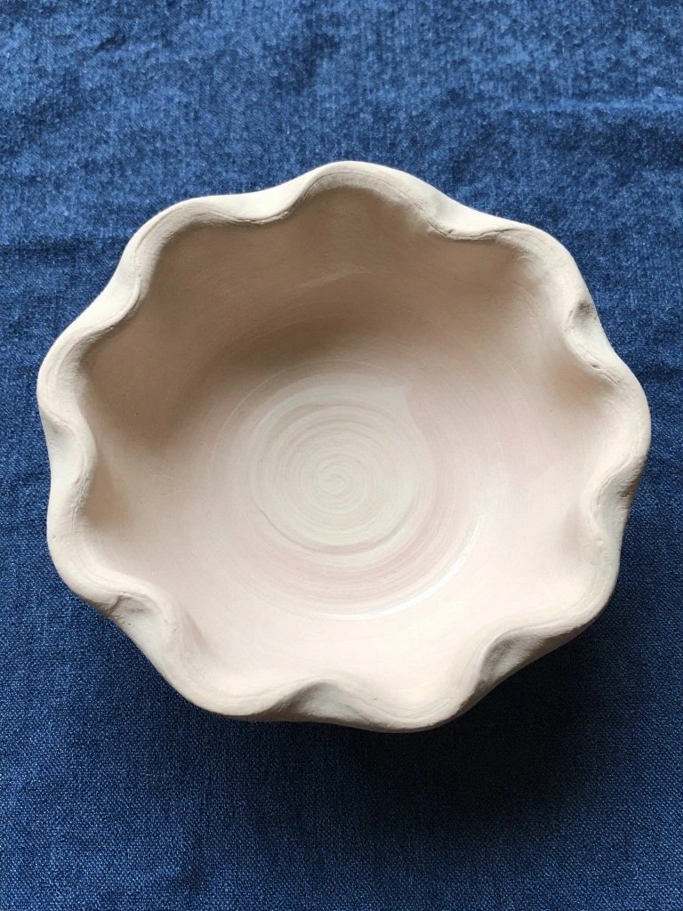 Small unfired clay bowl with wavy scalloped rim and pale spiral interior on blue denim fabric.