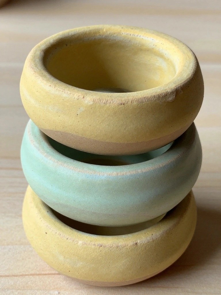 Three small handmade ceramic bowls glazed yellow and teal stacked on a wooden surface.