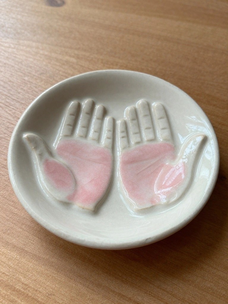 Small white ceramic dish molded as two facing hands with pink-glazed palms sitting on a wooden table.