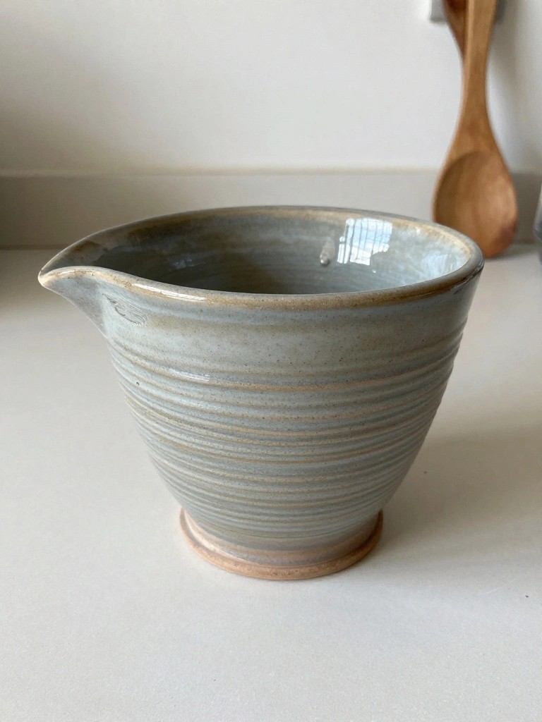 Handmade light gray ceramic bowl with vertical ridges, pouring spout, and footed base on a kitchen counter next to a wooden spoon.