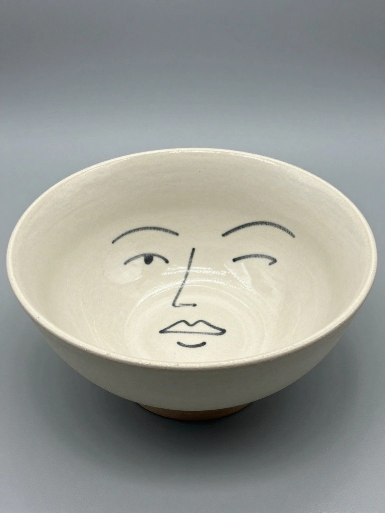 A white footed ceramic bowl with a black line-drawn face of arched eyebrows, eyes, and mouth on the interior.