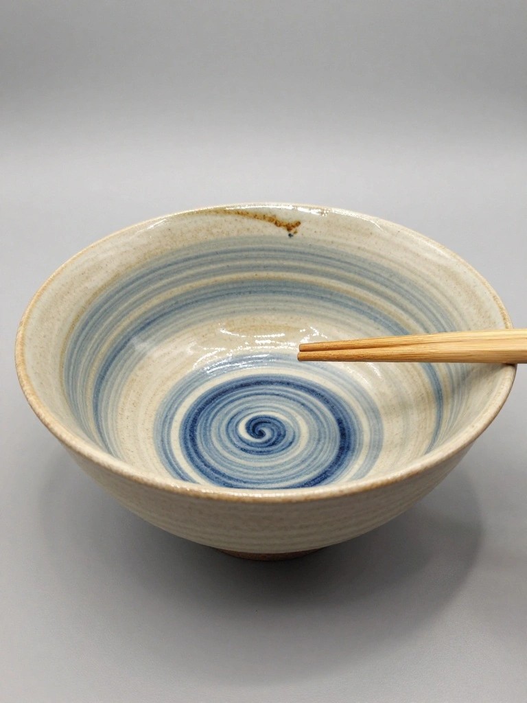 Wide handmade ceramic bowl with swirling blue glaze inside, beige exterior, and chopsticks resting across the rim.