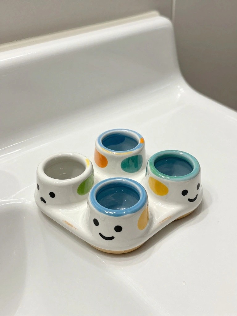 White ceramic tray with four small cups in blue, orange, green, and teal rims forming smiley faces, placed on a bathroom counter with blue liquid in the cups.