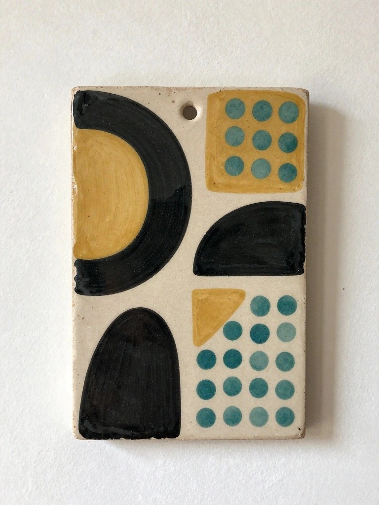 Rectangular handmade ceramic tile with abstract black geometric shapes, yellow curves, teal dotted patterns, and a top hanging hole on a light clay base.