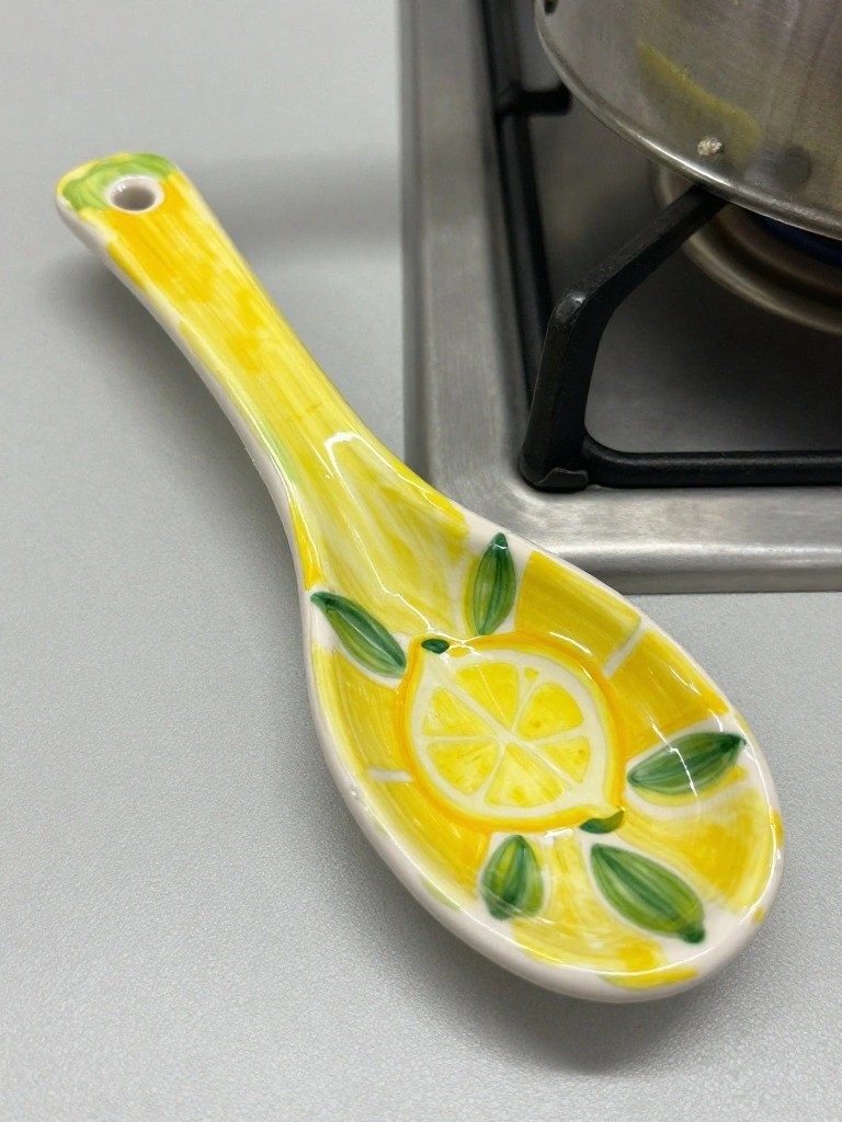 Yellow ceramic spoon with painted lemon slice in the bowl and green leaves on the handle sits on a kitchen stove next to a pot.