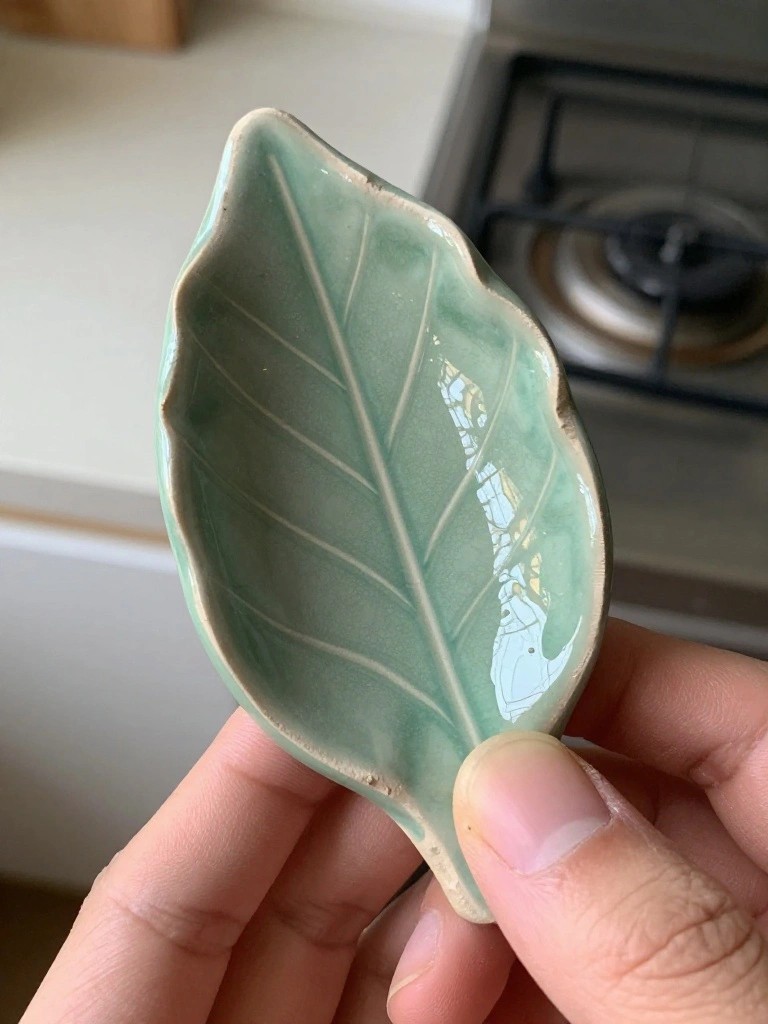 A small pale green glazed ceramic dish shaped like a leaf with textured veins, held in a hand on a kitchen counter near a stove.
