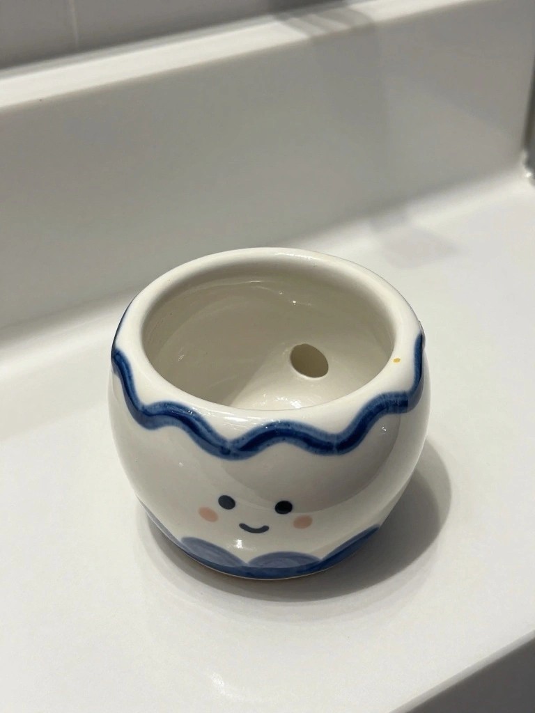 A small round white ceramic planter with blue wavy edges and a cute smiling face sits on a white tiled bathroom surface.