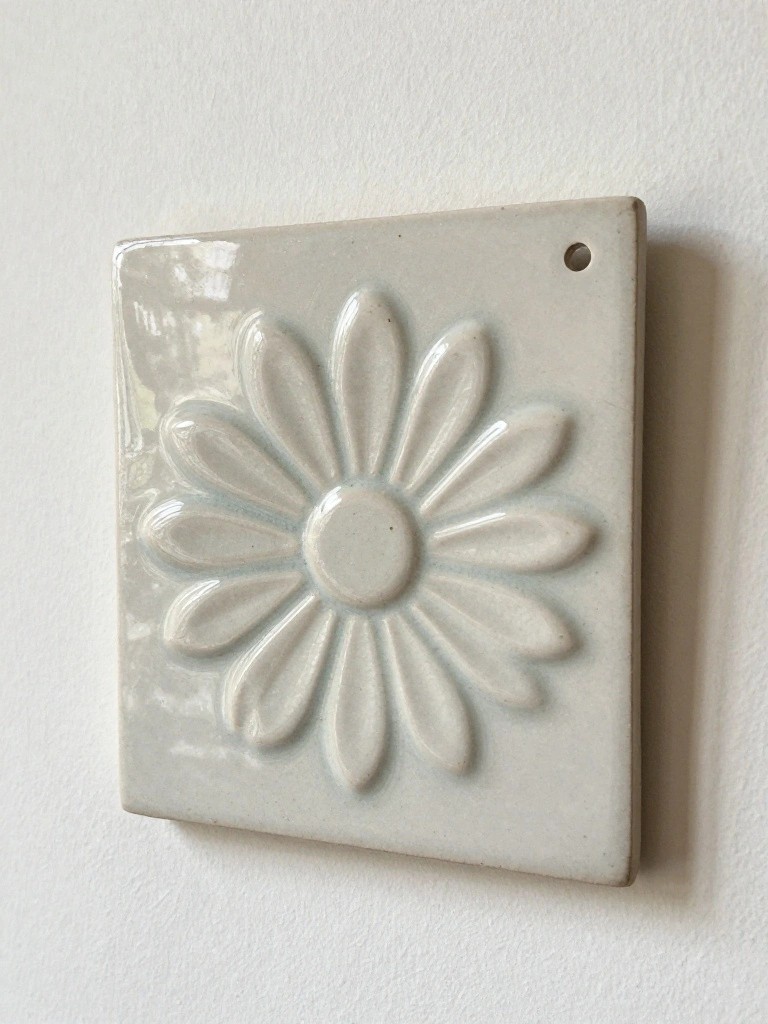 Square white glazed ceramic tile with raised daisy relief design and top hanging hole, mounted on white wall.