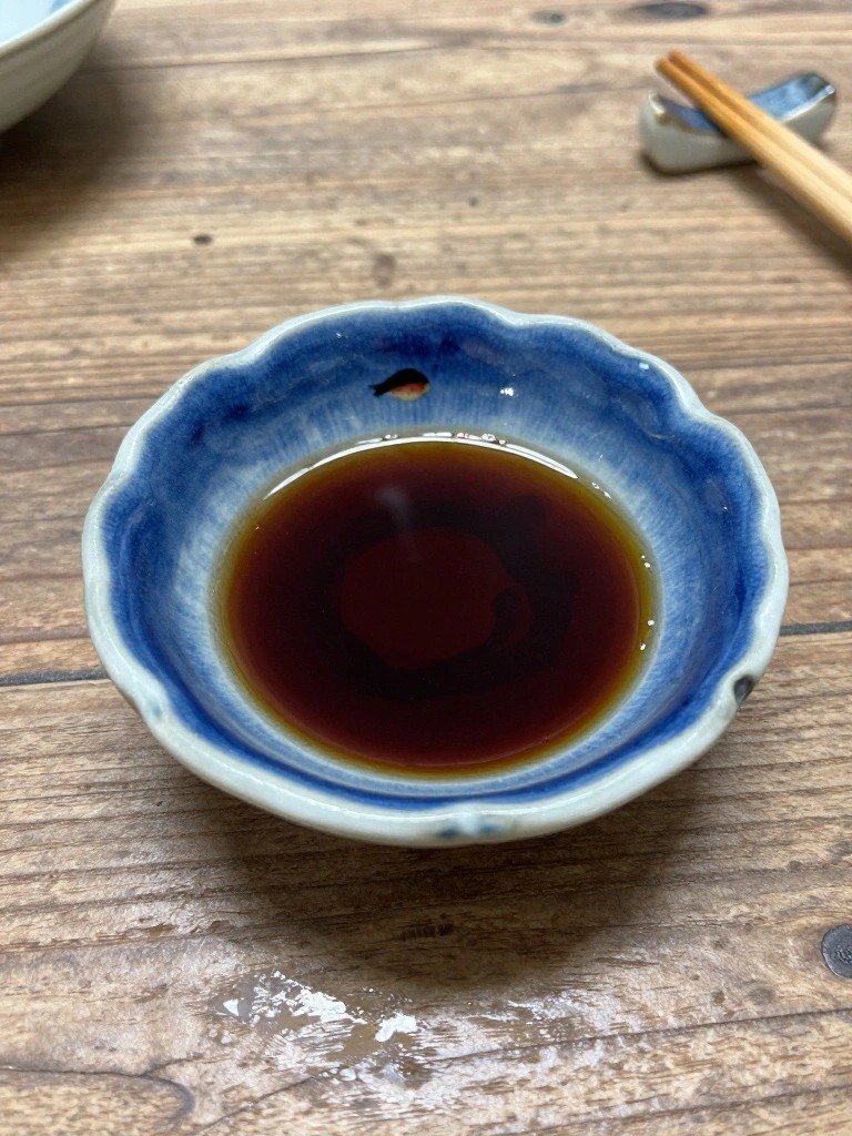Small blue ceramic bowl with scalloped edges and dark liquid inside sits on a wooden table with chopsticks nearby.