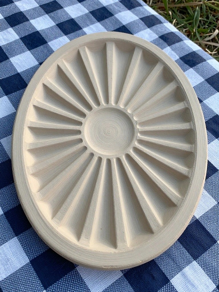 Unglazed oval clay tray with 18 carved radial ridges fanning from a central circle, on a blue checkered picnic tablecloth outdoors.