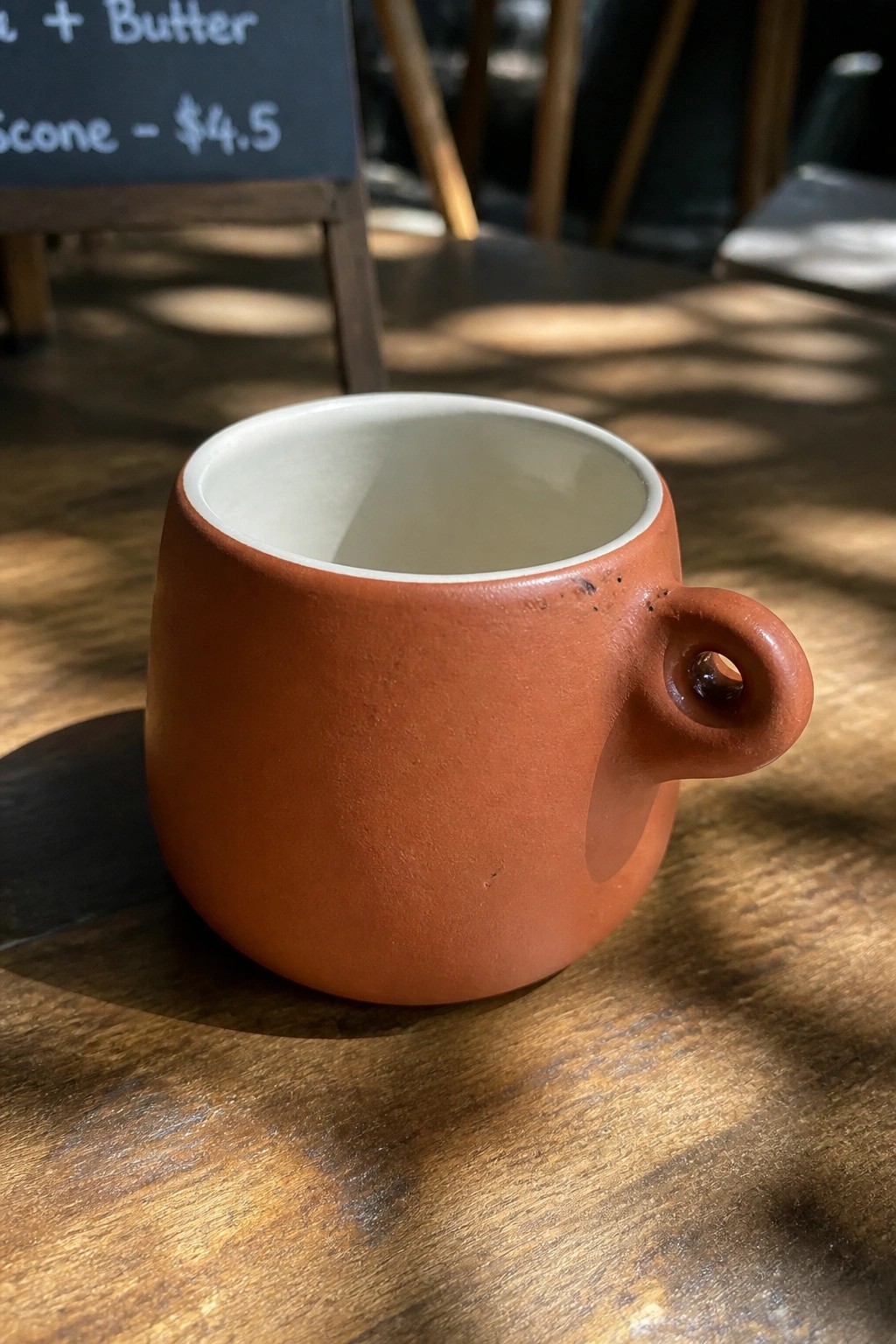 Terracotta cup with holed handle on sunlit wooden cafe table
