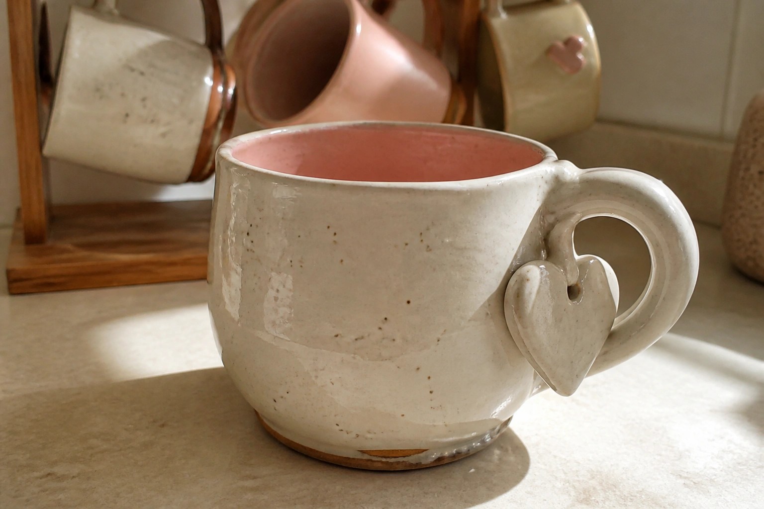 Cream ceramic mug with pink interior and heart accent on sunny surface.