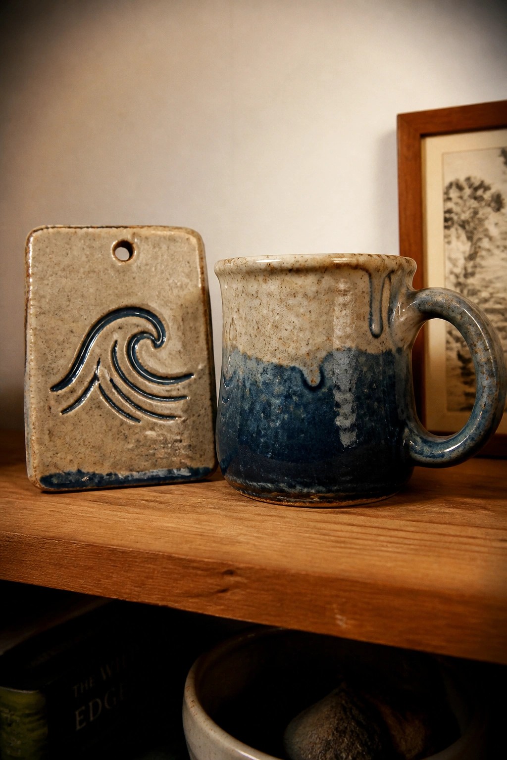 Ceramic mug with beige and blue glaze next to a rectangular tile carved with a wave on a wooden shelf.