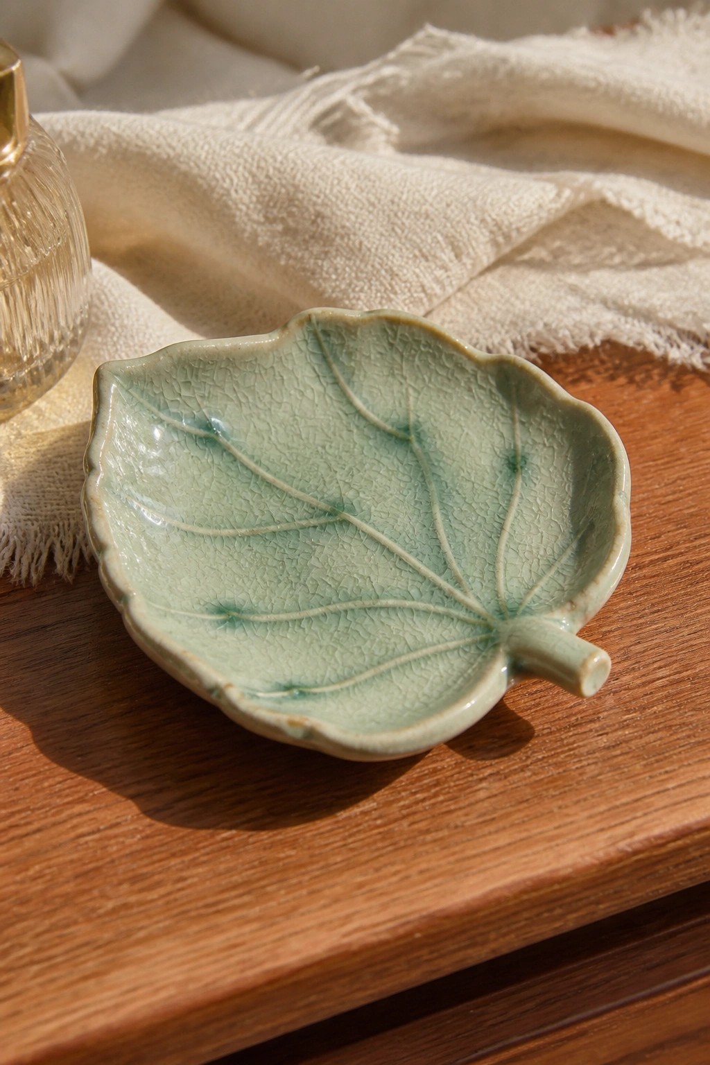 Crackled green ceramic leaf dish on wooden table with white cloth and glass bottle