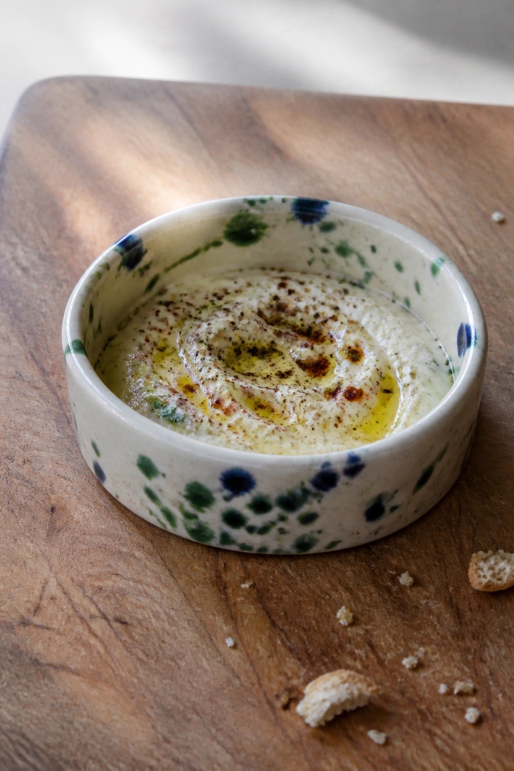 Creamy hummus swirled with oil and spices in a speckled ceramic bowl on wood