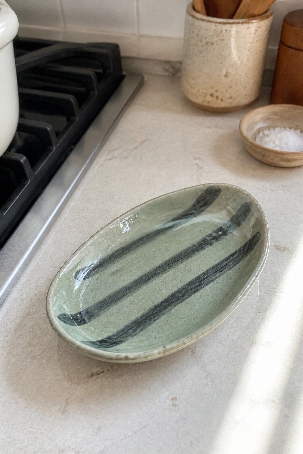 A small oval ceramic dish with light green glaze and dark striped brush marks sits on a marble countertop.
