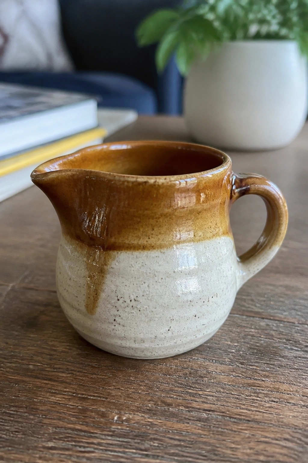 Small brown and cream ceramic pitcher on wooden table with books and plant.