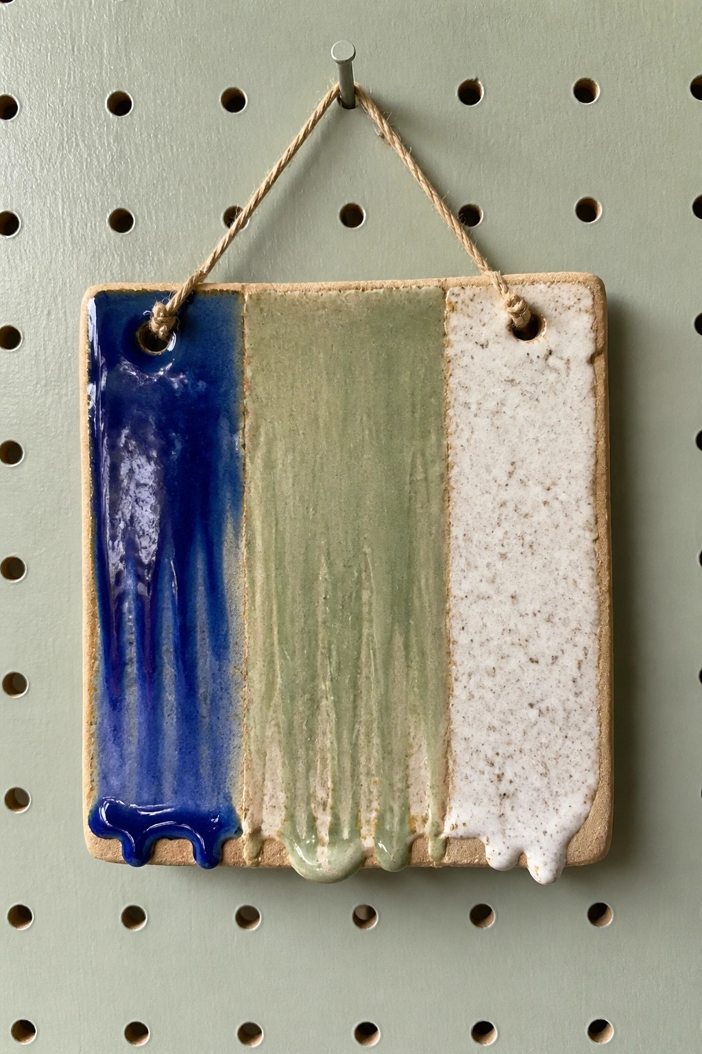 Ceramic tile with dripping blue, green, and speckled white glazes on pegboard.