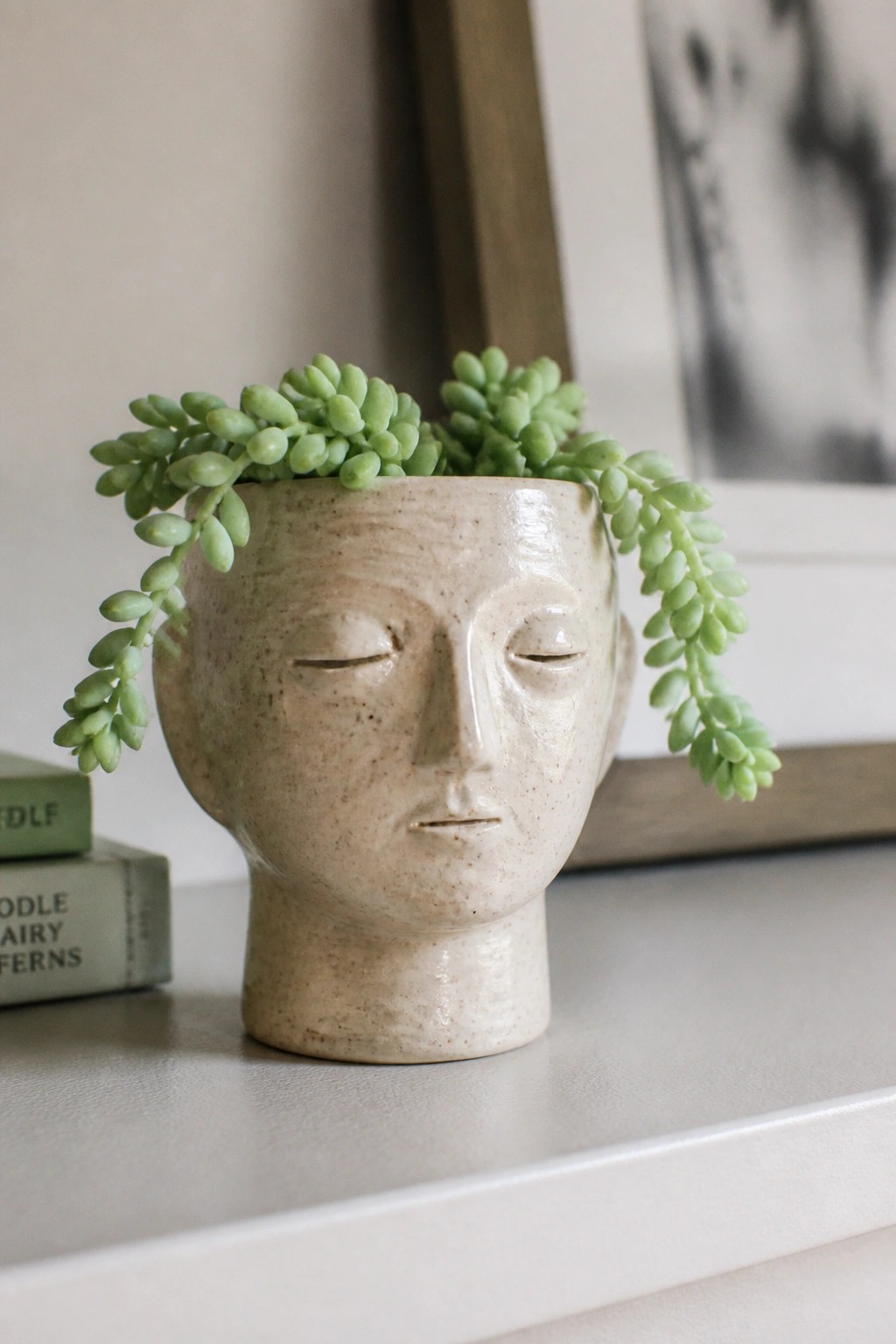 Ceramic head planter with closed eyes holding trailing green succulent on shelf.
