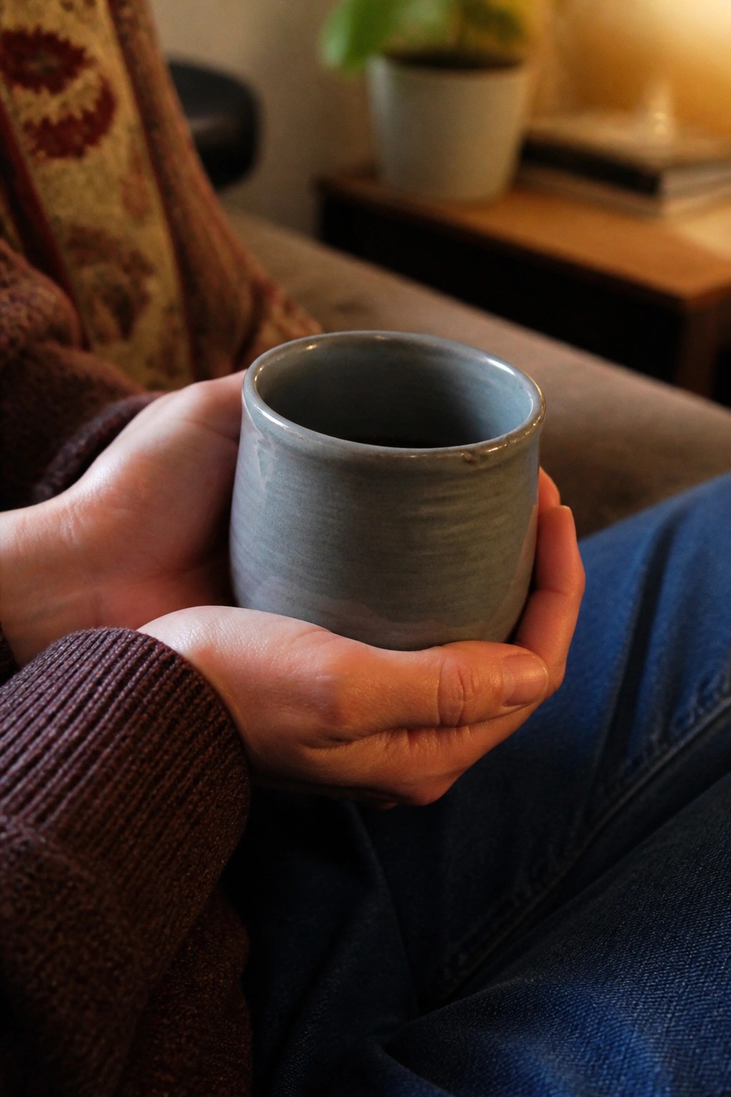Hands holding a blue ceramic mug, wearing brown sweater and blue jeans.