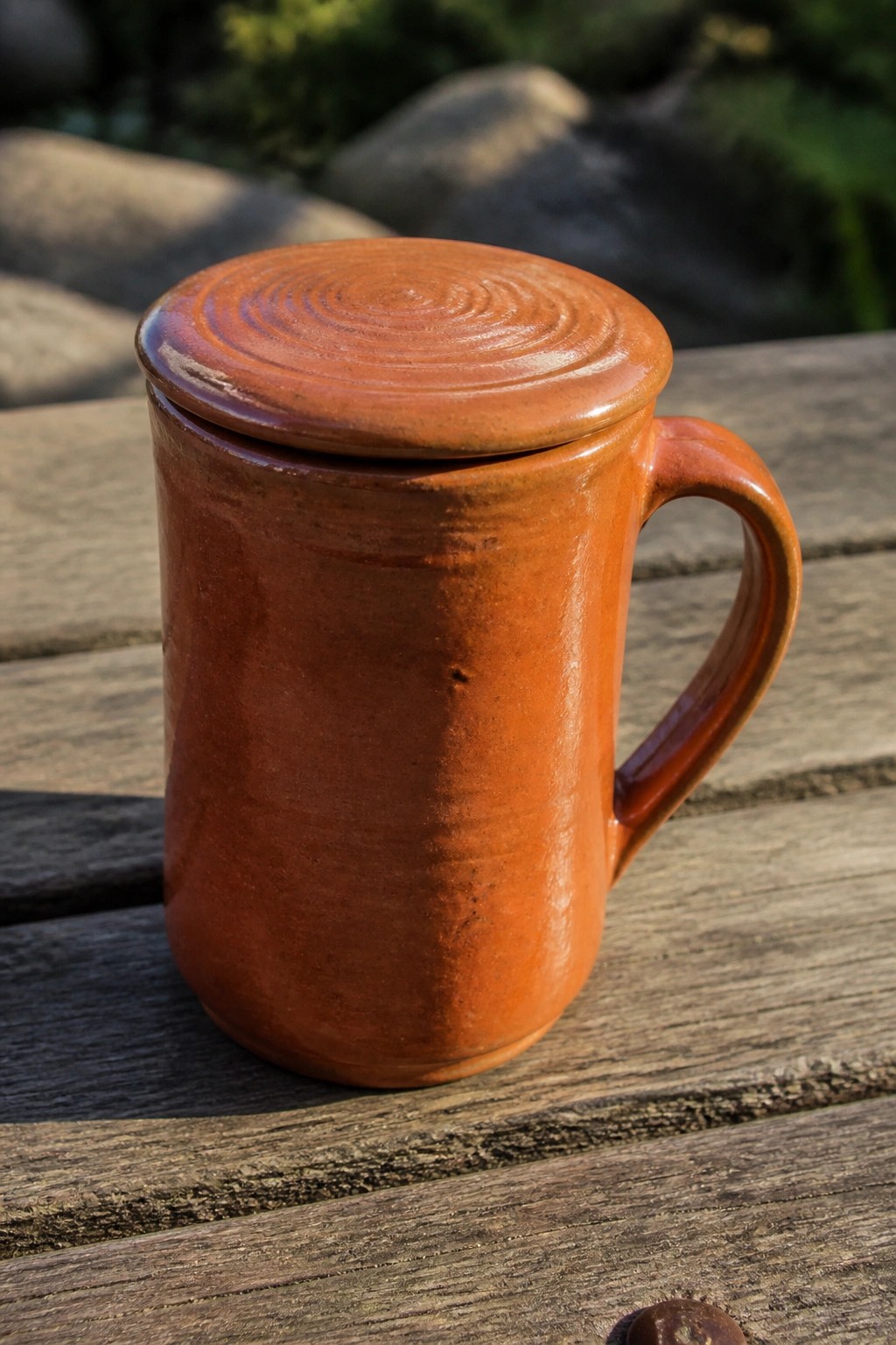 Glossy terracotta mug with spiral lid on sunlit wooden table outdoors