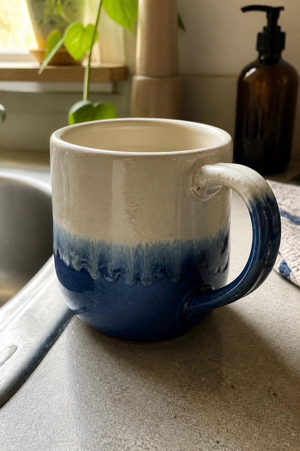 A ceramic mug with white glaze on the top half and blue glaze on the bottom half sits on a gray countertop near a sink.