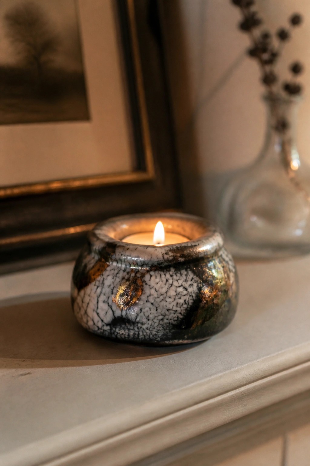 Lit candle in crackled multicolored ceramic holder on mantel with framed art nearby.
