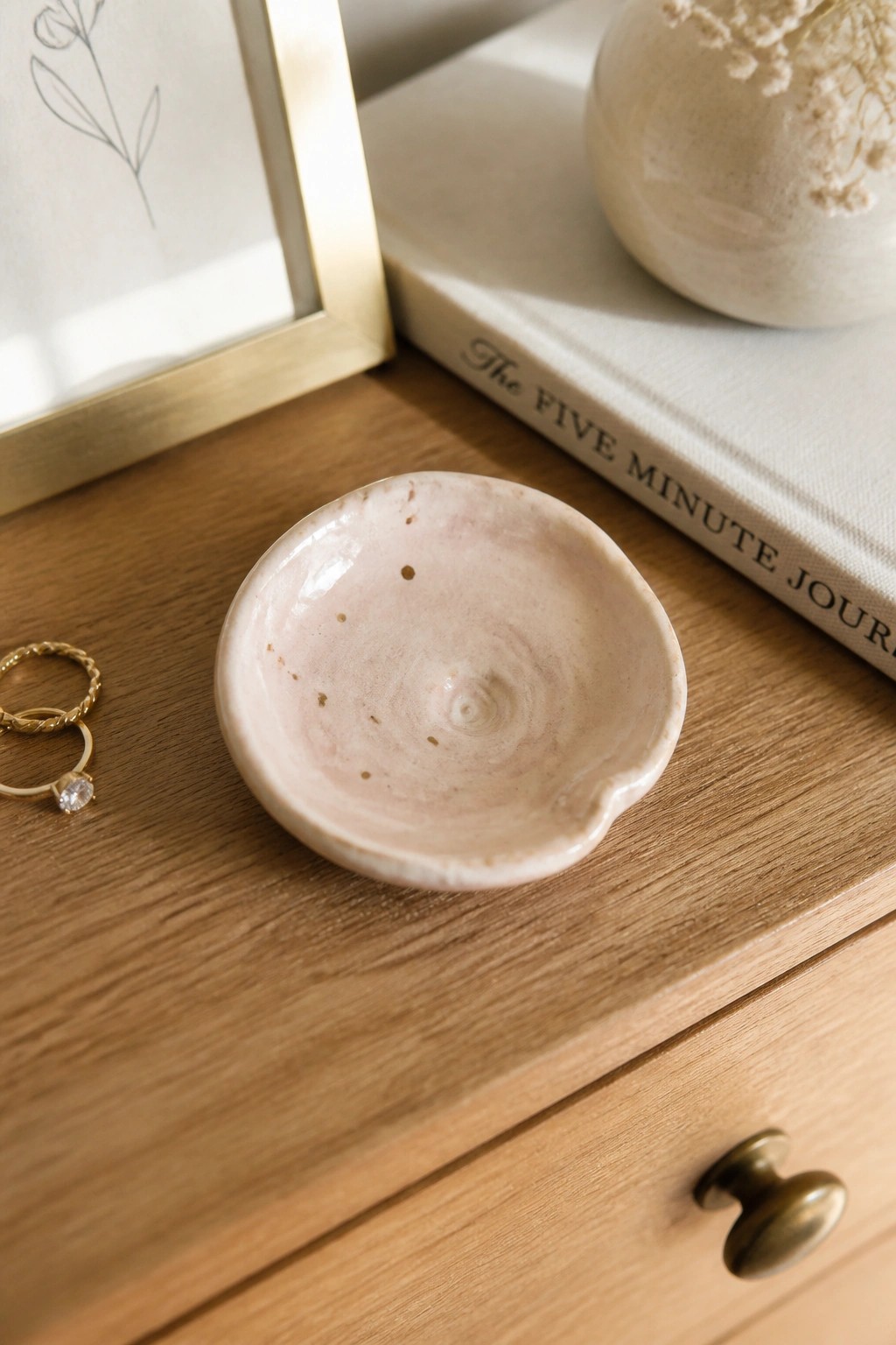 A small pink ceramic dish with gold speckles sits on a wooden surface next to gold rings, a book, and a vase.