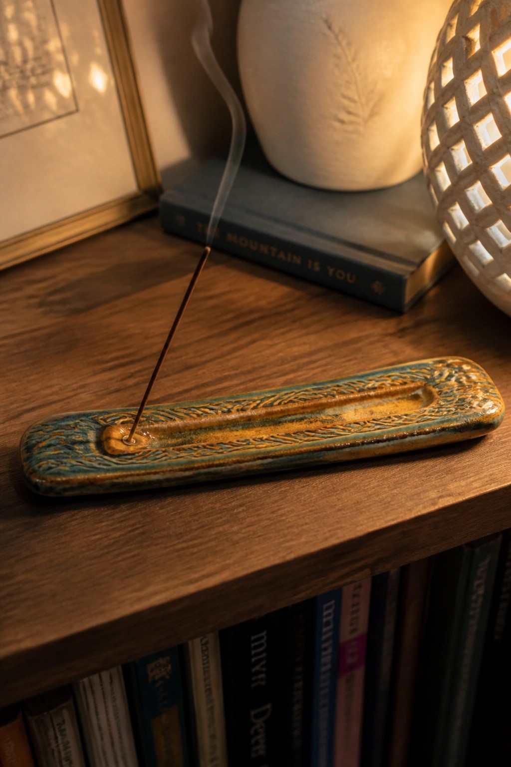Burning incense stick in ornate turquoise ceramic holder on wooden shelf with books
