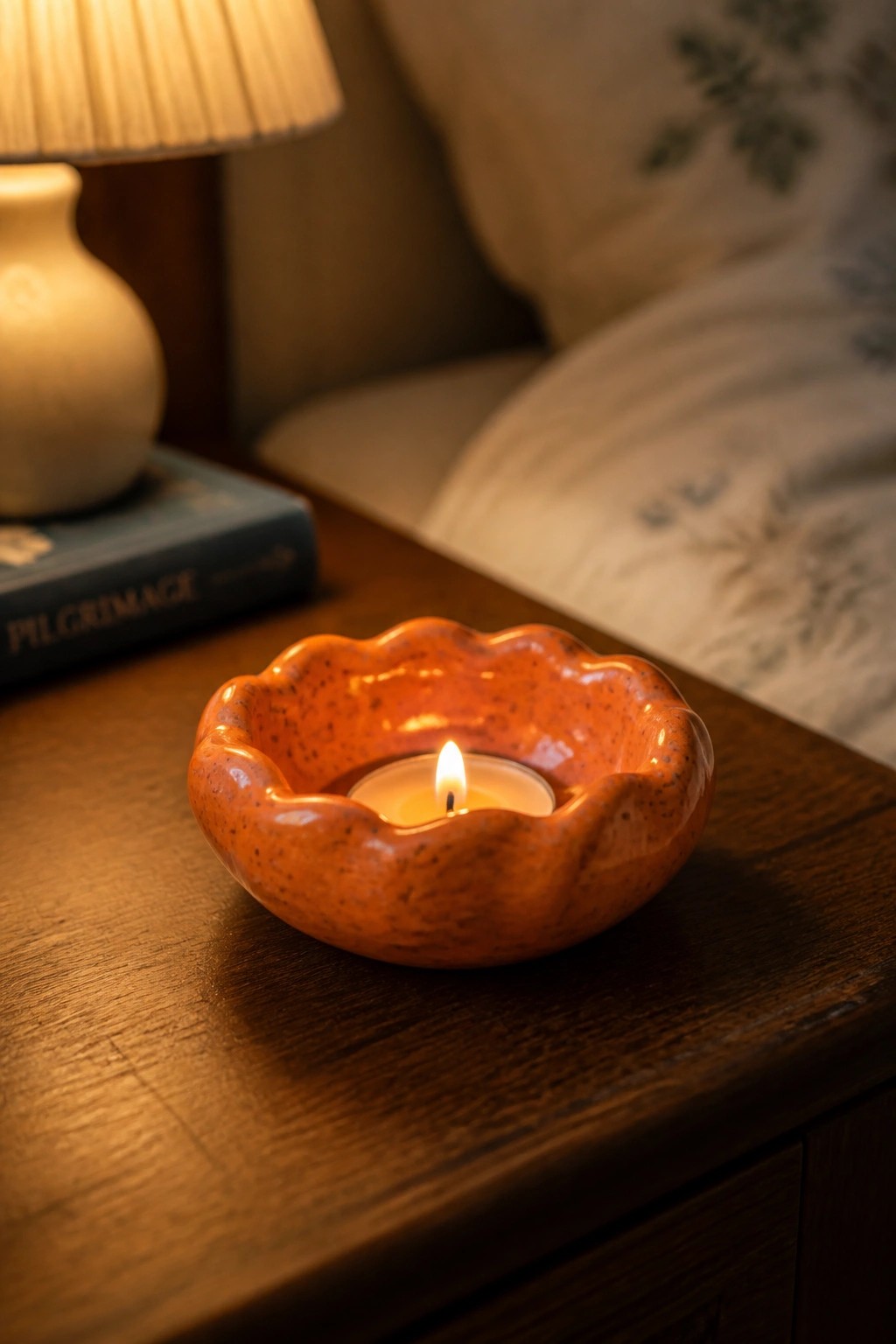 Lit candle in orange speckled ceramic holder on wooden table beside book and lamp