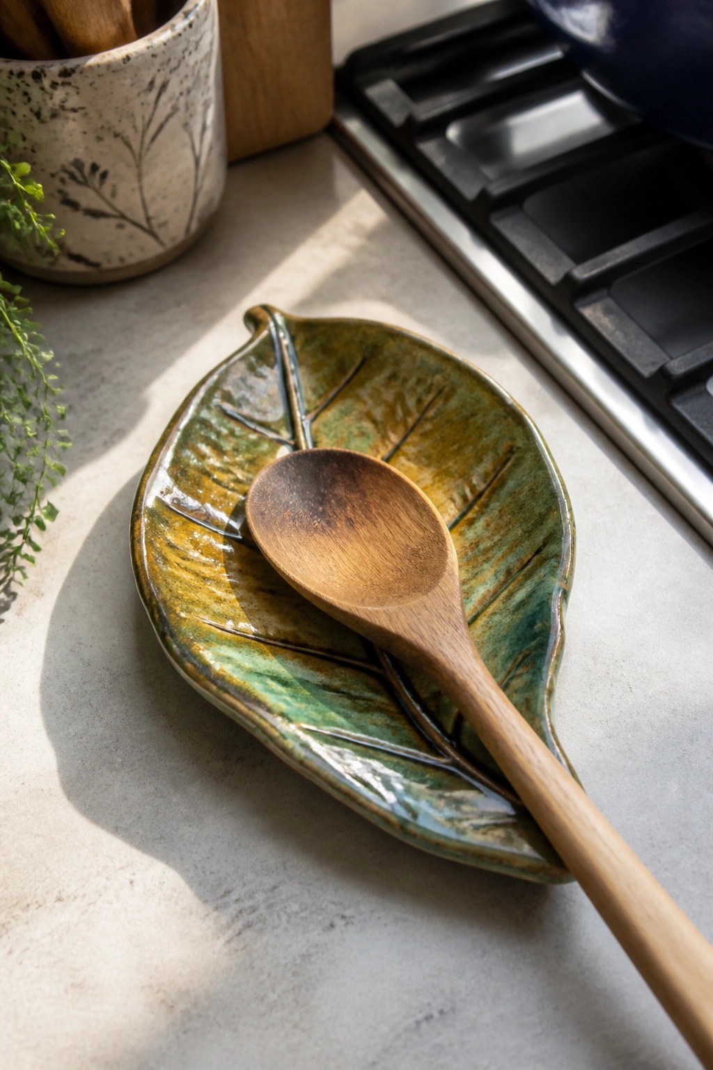 A wooden spoon rests on a green and brown glazed ceramic leaf-shaped dish on a kitchen counter.