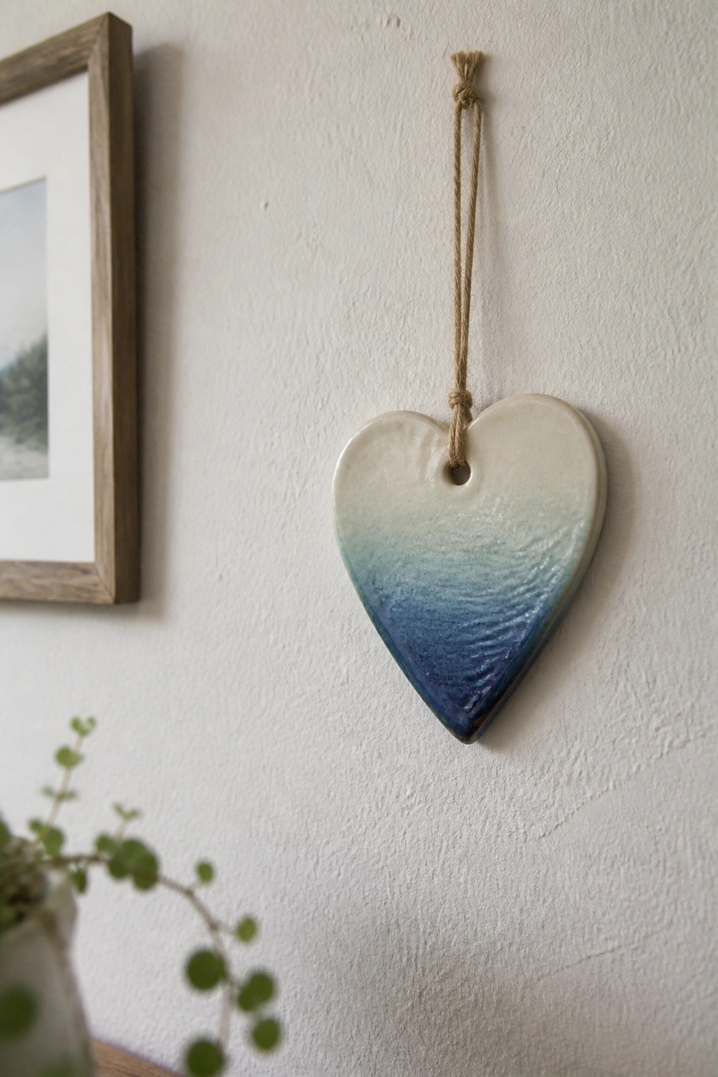 A heart-shaped ceramic ornament with blue gradient glaze hangs from twine on a white wall next to a wooden picture frame.