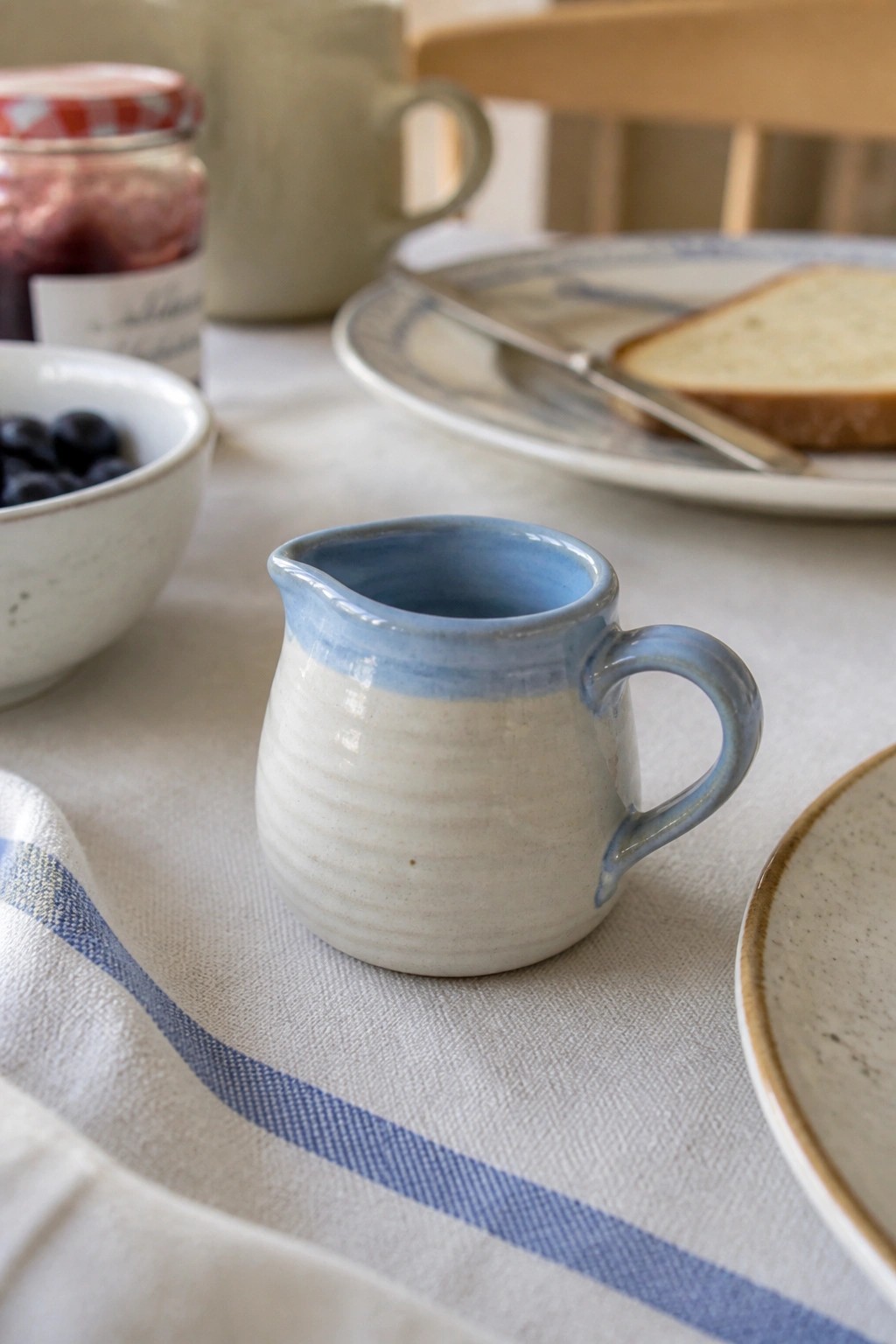 Small white-and-blue ceramic pitcher on table with jam jar, blueberries, and bread.