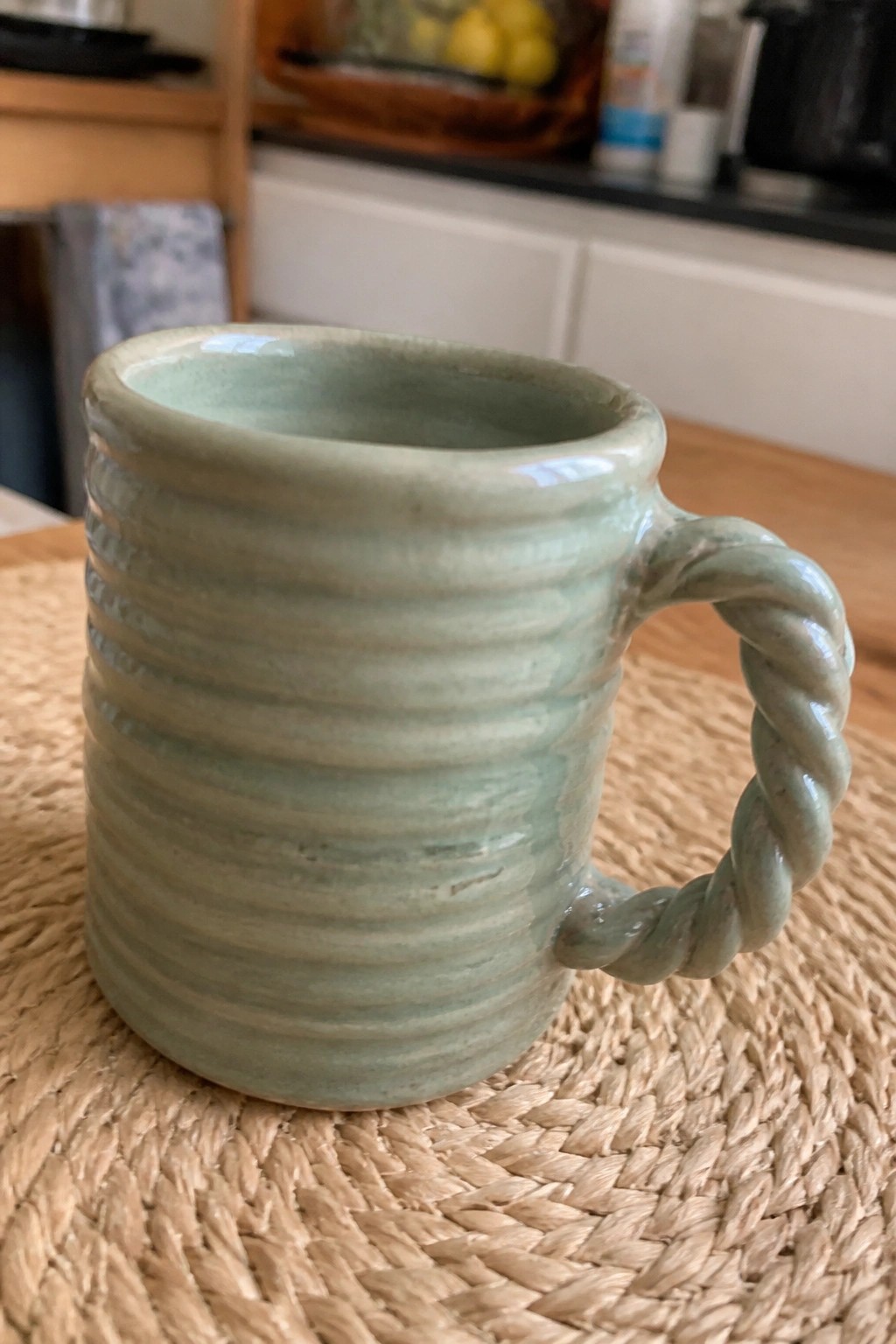 A light green ceramic mug with horizontal ridges and a twisted rope handle sits on a woven placemat.