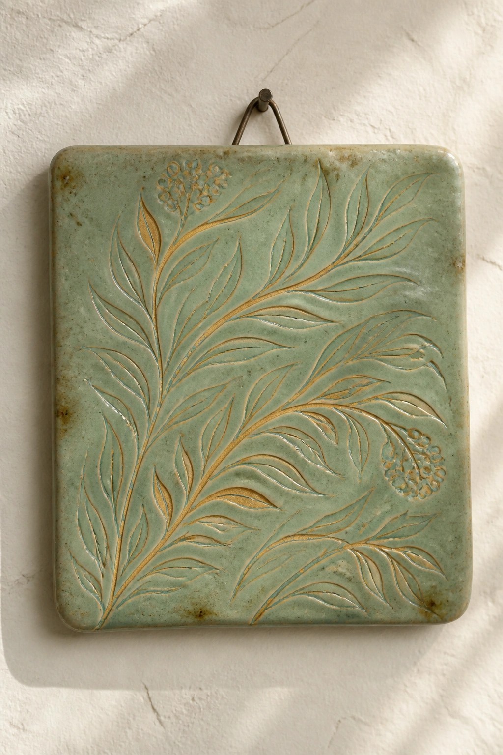 A square green ceramic tile with embossed leaf and flower designs hangs on a white wall by a metal wire.