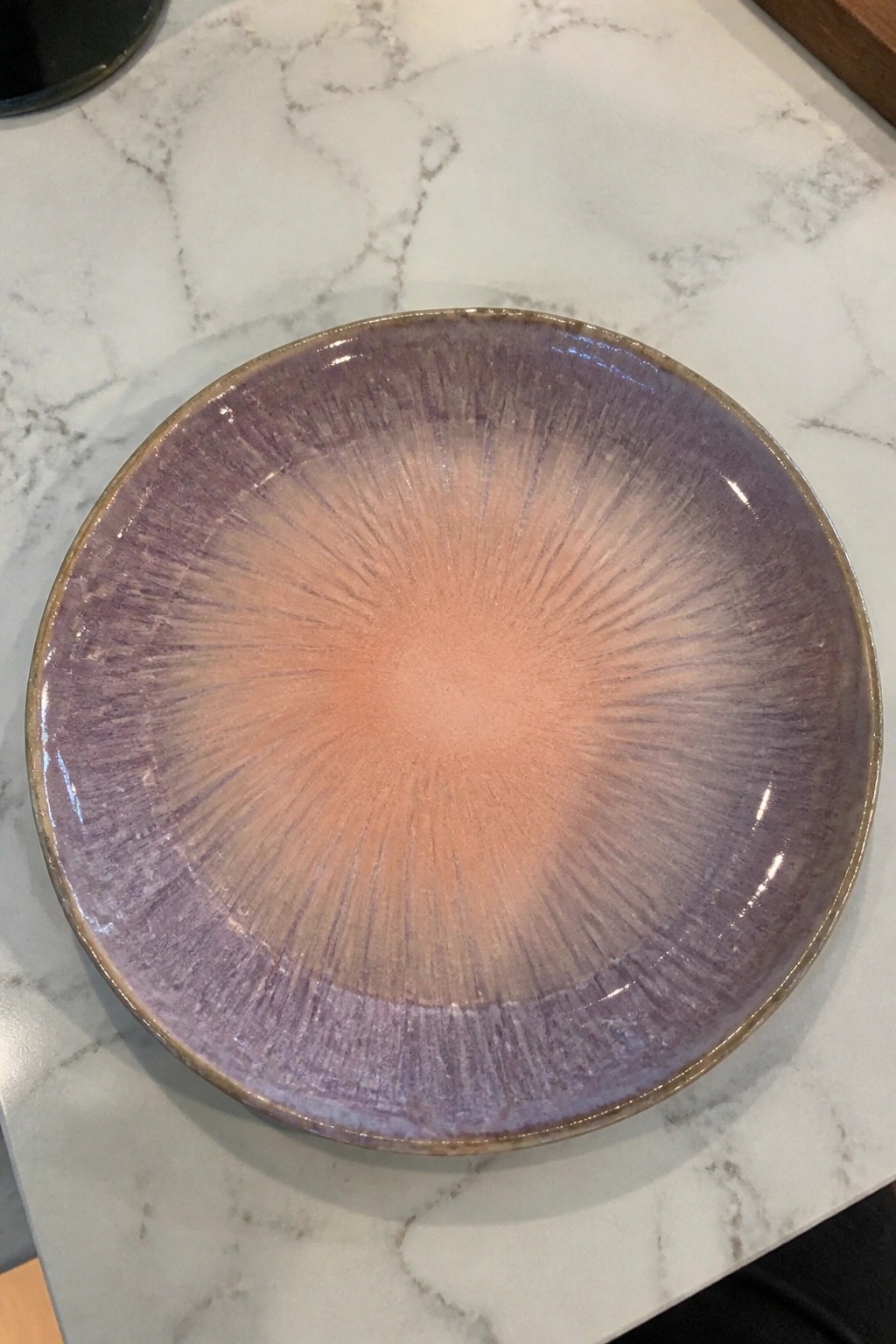 A ceramic shallow bowl with purple to peach radial glaze on a white marble surface.