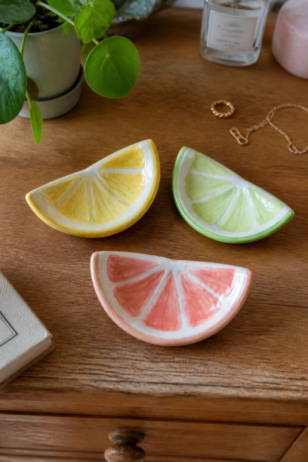 Three ceramic citrus slices—lemon, lime, grapefruit—on a wooden table with jewelry and plant.