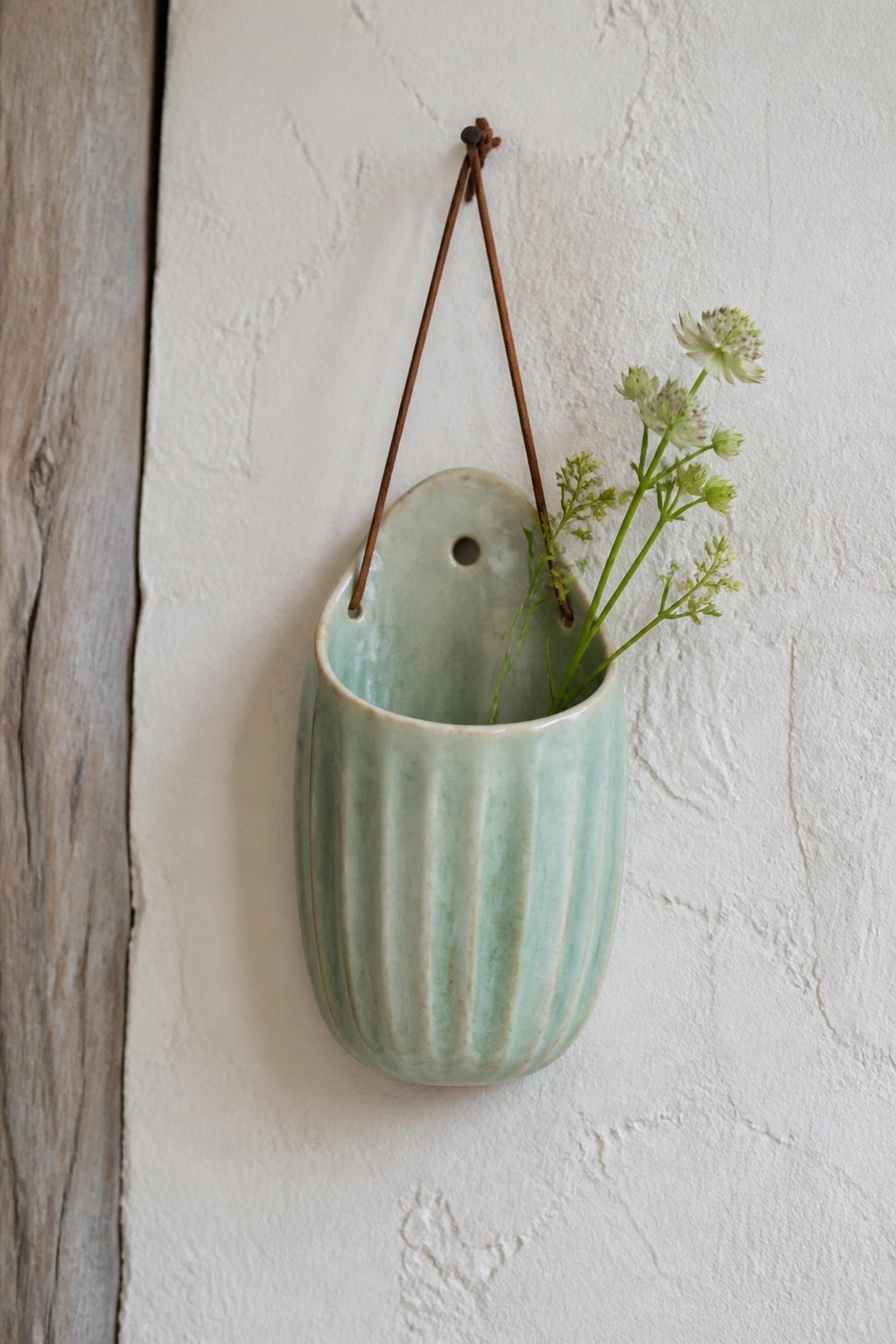 Light green ribbed ceramic vase hanging by cord on white wall with flowers