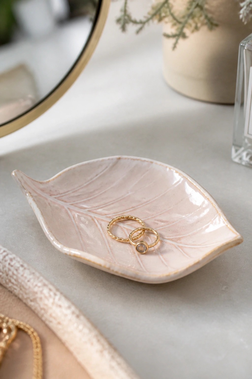 A pink ceramic leaf-shaped dish with gold edges holding two gold rings on a marble surface.