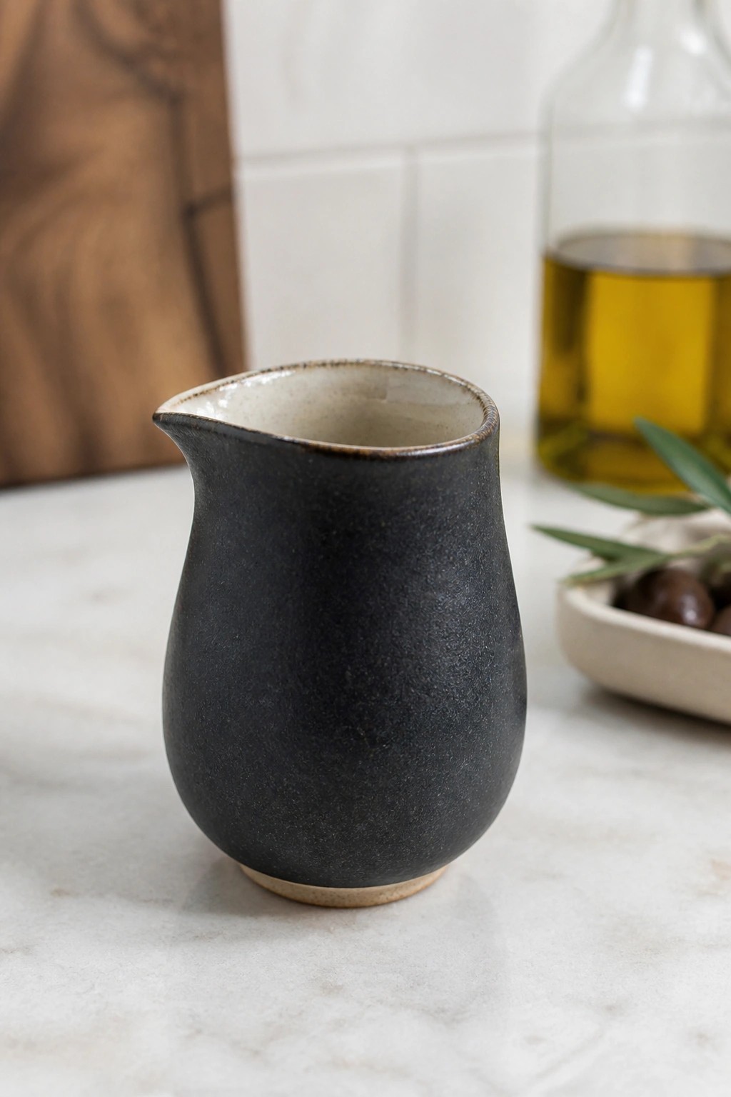 Black ceramic pitcher on marble counter with olive oil bottle and olives nearby