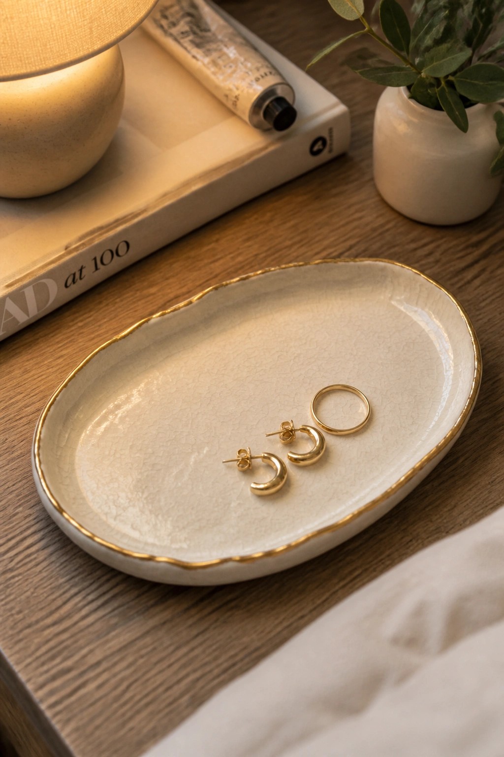 A white oval ceramic tray with a gold rim holding gold hoop earrings and a ring on a wooden surface.