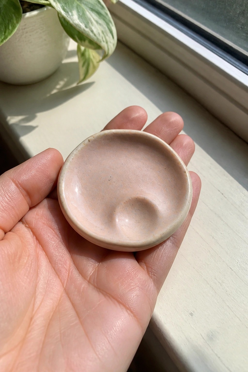 A small round ceramic dish with pink speckled glaze held in a hand on a windowsill.