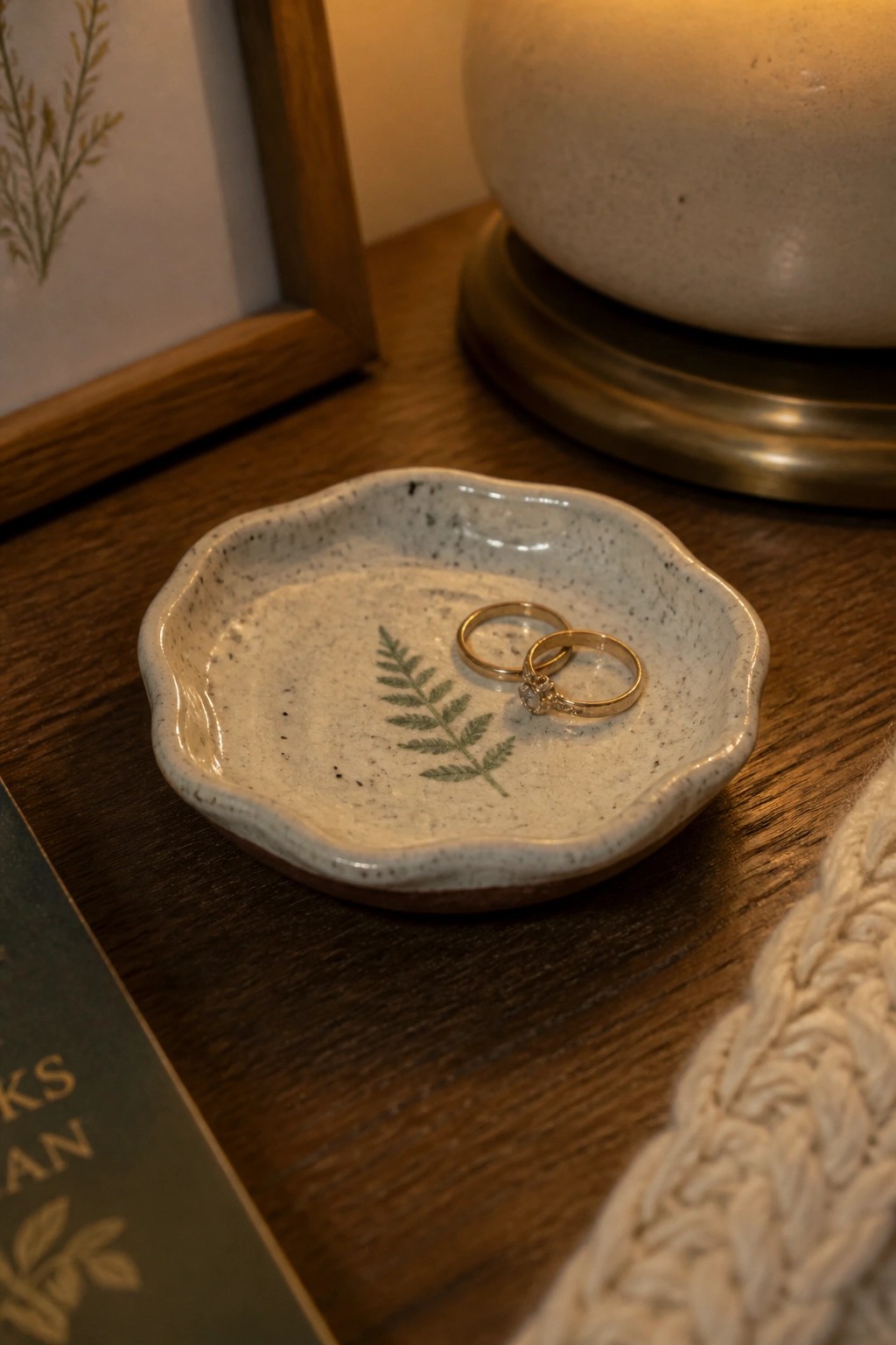 A small ceramic dish with scalloped edges and a green fern design holds two gold rings on a wooden table.