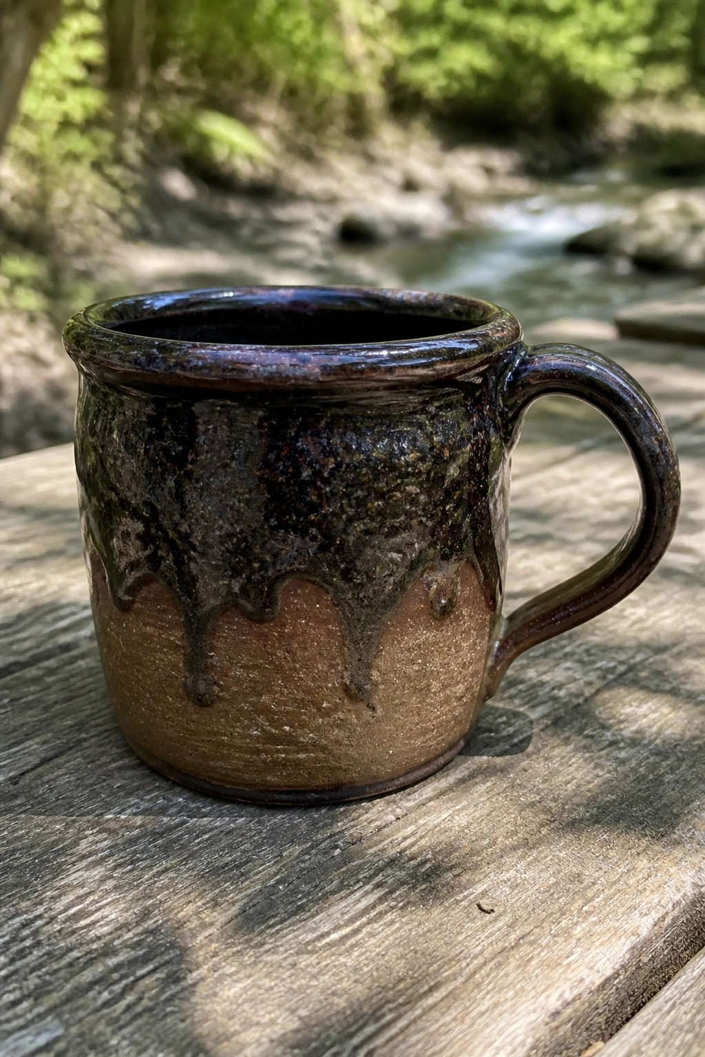 Ceramic mug with dripping dark glaze on wooden table beside forest stream